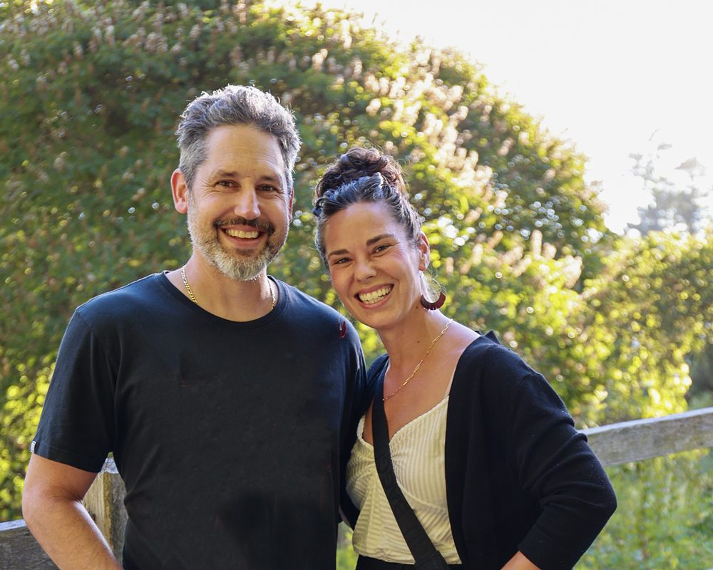 A caucasian man and woman look at the camera and are smiling, with a background of green leaves