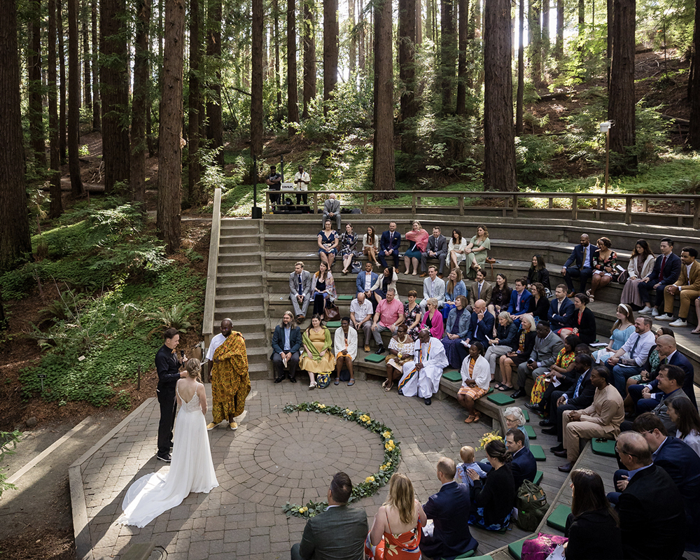 Many people seated on a tiered area watching two people get married with tall trees in the background