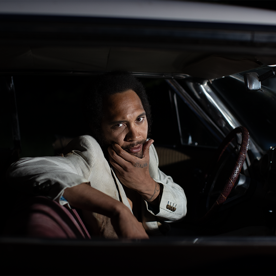 A black man with his hand on his chin is sitting in a car, looking at the camera