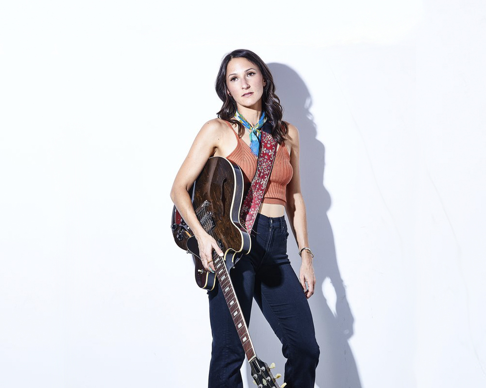 One woman stands against a white wall holding a guitar