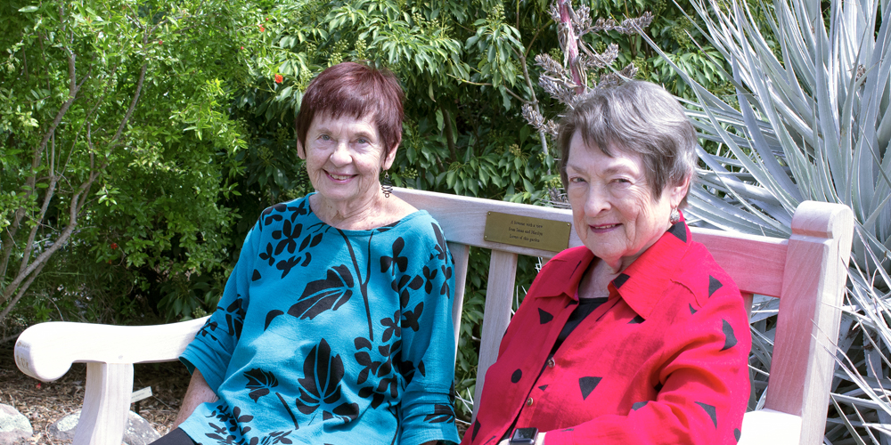 2 smiling women sit on a bench in a garden