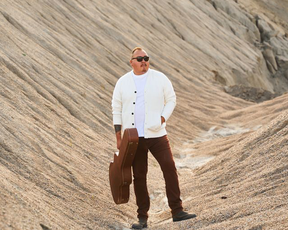 A man wearing sunglasses stands on rock and holds a guitar