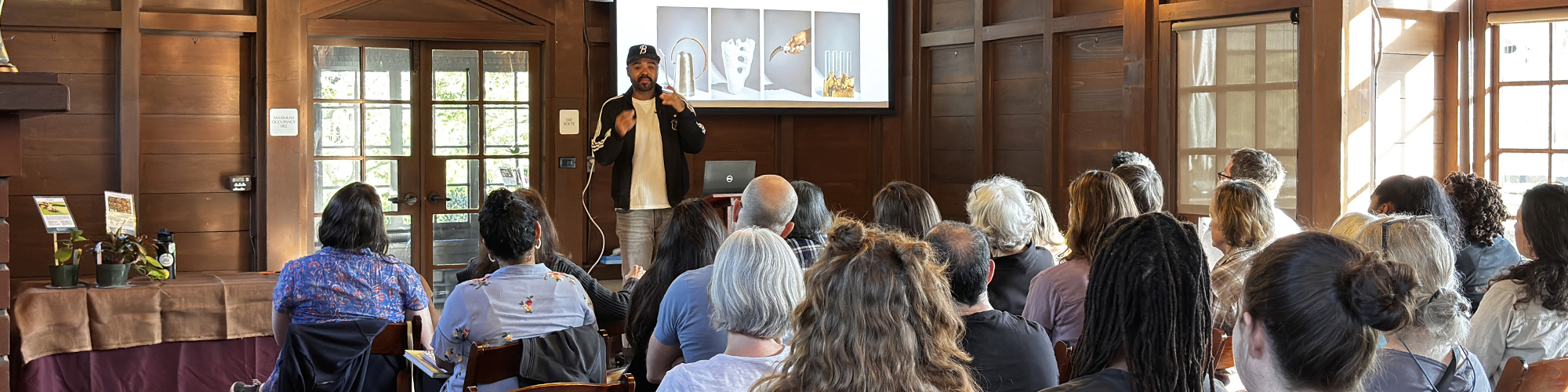 A group of people sit in a large room and watch a black man talk