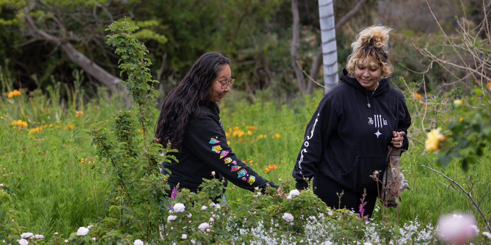 Two smiling young women stroll through a garden looking at flowers