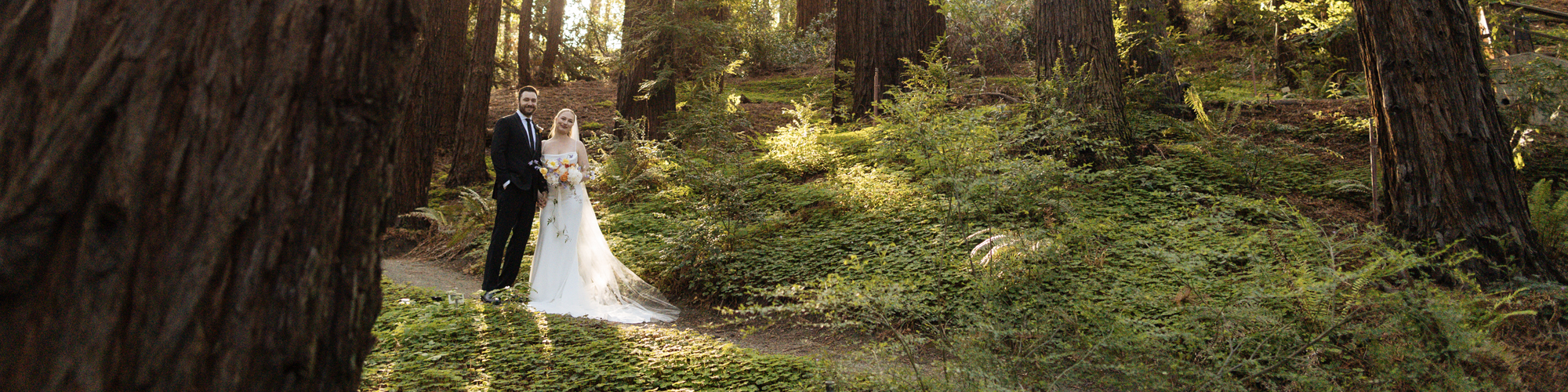 A bride and groom stand close together in a forest of trees