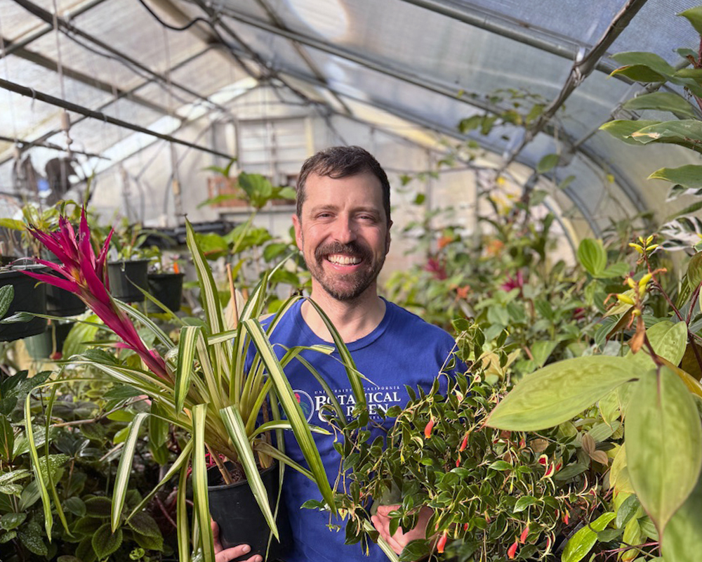 A smiling man is surrounded by houseplants in a greenhouse